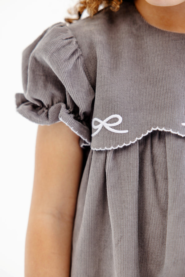 Here’s a rewritten version using your product data: A close-up showcases a girl wearing The Beaufort Bonnet Company’s McFarland Frock in Grantley Gray Corduroy with Worth Avenue White. The gray corduroy dress features puffed sleeves and delicate white embroidered detailing accenting the scalloped edge near the shoulder, set against a white background. This charming fall dress for girls is adorned with thoughtful accents that highlight its timeless appeal.
