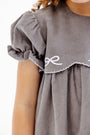 Here’s a rewritten version using your product data: A close-up showcases a girl wearing The Beaufort Bonnet Company’s McFarland Frock in Grantley Gray Corduroy with Worth Avenue White. The gray corduroy dress features puffed sleeves and delicate white embroidered detailing accenting the scalloped edge near the shoulder, set against a white background. This charming fall dress for girls is adorned with thoughtful accents that highlight its timeless appeal.