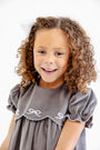 A smiling young girl with curly hair stands against a white background wearing The Beaufort Bonnet Company’s McFarland Frock in Grantley Gray Corduroy with Worth Avenue White, paired with a white embroidered bow in her hair.