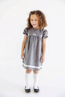 A young girl with curly hair, wearing The Beaufort Bonnet Company’s McFarland Frock in Grantley Gray Corduroy with Worth Avenue White, white knee-high socks, and black shoes, stands smiling against a plain white background.