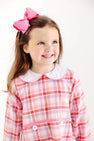 A young girl with freckles wears a long-sleeved Lindy’s Lunch Dress - Broadway Play Plaid with Worth Avenue White, featuring a white collar and a large pink hair bow.