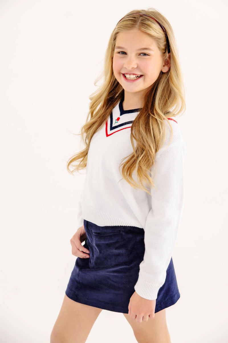 A girl wearing a white V-neck sweater with red and navy trim and a Stevi Skirt - Nantucket Navy Corduroy with an adjustable waistband.