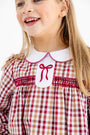 A smiling young girl with long blonde hair wears The Beaufort Bonnet Company Long Sleeve Mary Dal Dress in Easton Pond Plaid with Worth Avenue White—a charming girls’ autumn dress featuring a classic plaid pattern in red, brown, and white, finished with a crisp white collar and sweet red bow detail.