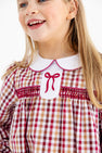 A smiling young girl with long blonde hair wears The Beaufort Bonnet Company Long Sleeve Mary Dal Dress in Easton Pond Plaid with Worth Avenue White—a charming girls’ autumn dress featuring a classic plaid pattern in red, brown, and white, finished with a crisp white collar and sweet red bow detail.