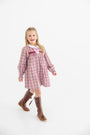 A young girl with long blonde hair, smiling with her mouth open, walks forward against a plain white background wearing The Beaufort Bonnet Company’s Long Sleeve Mary Dal Dress in Easton Pond Plaid with Worth Avenue White, paired with brown knee-high boots.