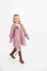 A young girl with long blonde hair, smiling with her mouth open, walks forward against a plain white background wearing The Beaufort Bonnet Company’s Long Sleeve Mary Dal Dress in Easton Pond Plaid with Worth Avenue White, paired with brown knee-high boots.
