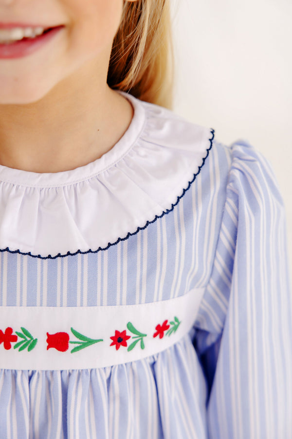 A child wearing a Long Sleeve Dollie Mae Dress - Park City Periwinkle Ticking Stripe & Worth Avenue White, featuring a white ruffled collar and an embroidered floral pattern across the chest.
