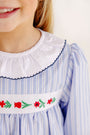 A child wearing a Long Sleeve Dollie Mae Dress - Park City Periwinkle Ticking Stripe & Worth Avenue White, featuring a white ruffled collar and an embroidered floral pattern across the chest.