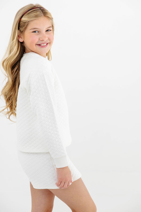 A young girl with long blonde hair smiles while wearing The Beaufort Bonnet Company’s Cassidy Comfy Crewneck in Quilted Palmetto Pearl with Nantucket Navy Bows, along with matching shorts, standing against a plain white background.