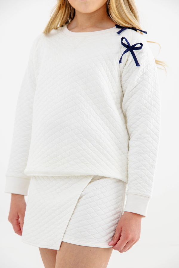 A girl is pictured wearing The Beaufort Bonnet Company’s Cassidy Comfy Crewneck in Quilted Palmetto Pearl with Nantucket Navy Bows, paired with a matching quilted white skirt. Only her lower face and upper body appear against a plain, light background.