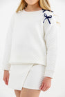 A girl is pictured wearing The Beaufort Bonnet Company’s Cassidy Comfy Crewneck in Quilted Palmetto Pearl with Nantucket Navy Bows, paired with a matching quilted white skirt. Only her lower face and upper body appear against a plain, light background.