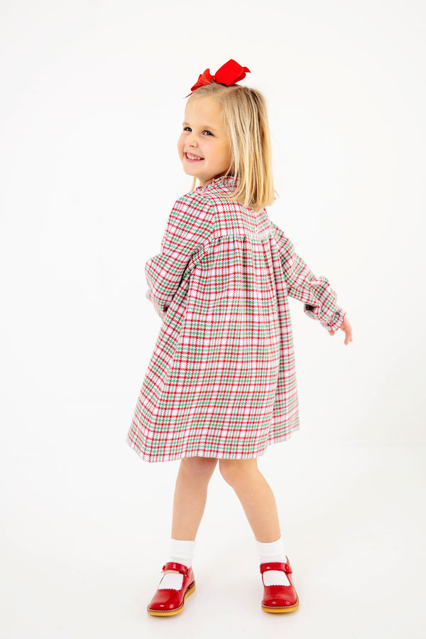 A young girl twirls and smiles against a white background, wearing The Beaufort Bonnet Company’s Long Sleeve Mary Baker Day Dress in Wicker Park Plaid with Richmond Red, paired with red shoes and a matching red bow in her hair.