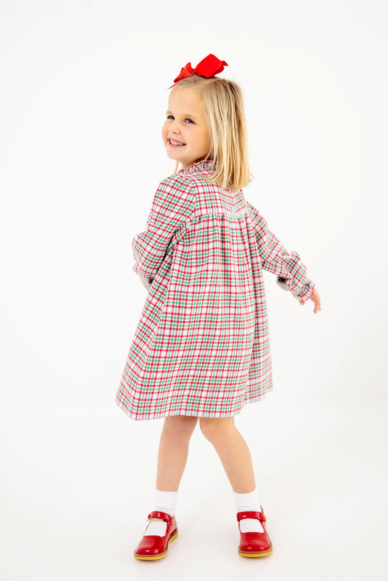 A young girl twirls and smiles against a white background, wearing The Beaufort Bonnet Company’s Long Sleeve Mary Baker Day Dress in Wicker Park Plaid with Richmond Red, paired with red shoes and a matching red bow in her hair.
