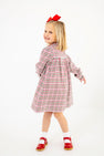 A young girl twirls and smiles against a white background, wearing The Beaufort Bonnet Company’s Long Sleeve Mary Baker Day Dress in Wicker Park Plaid with Richmond Red, paired with red shoes and a matching red bow in her hair.