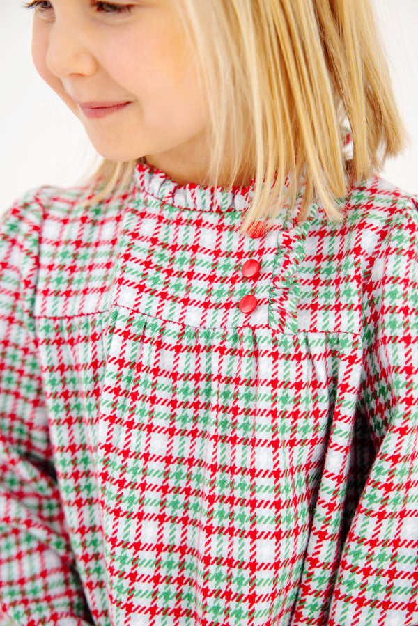 A smiling young child with blonde hair wears The Beaufort Bonnet Company’s Long Sleeve Mary Baker Day Dress in Wicker Park Plaid with Richmond Red—featuring three red buttons on the chest, it’s the perfect Christmas holiday dress.