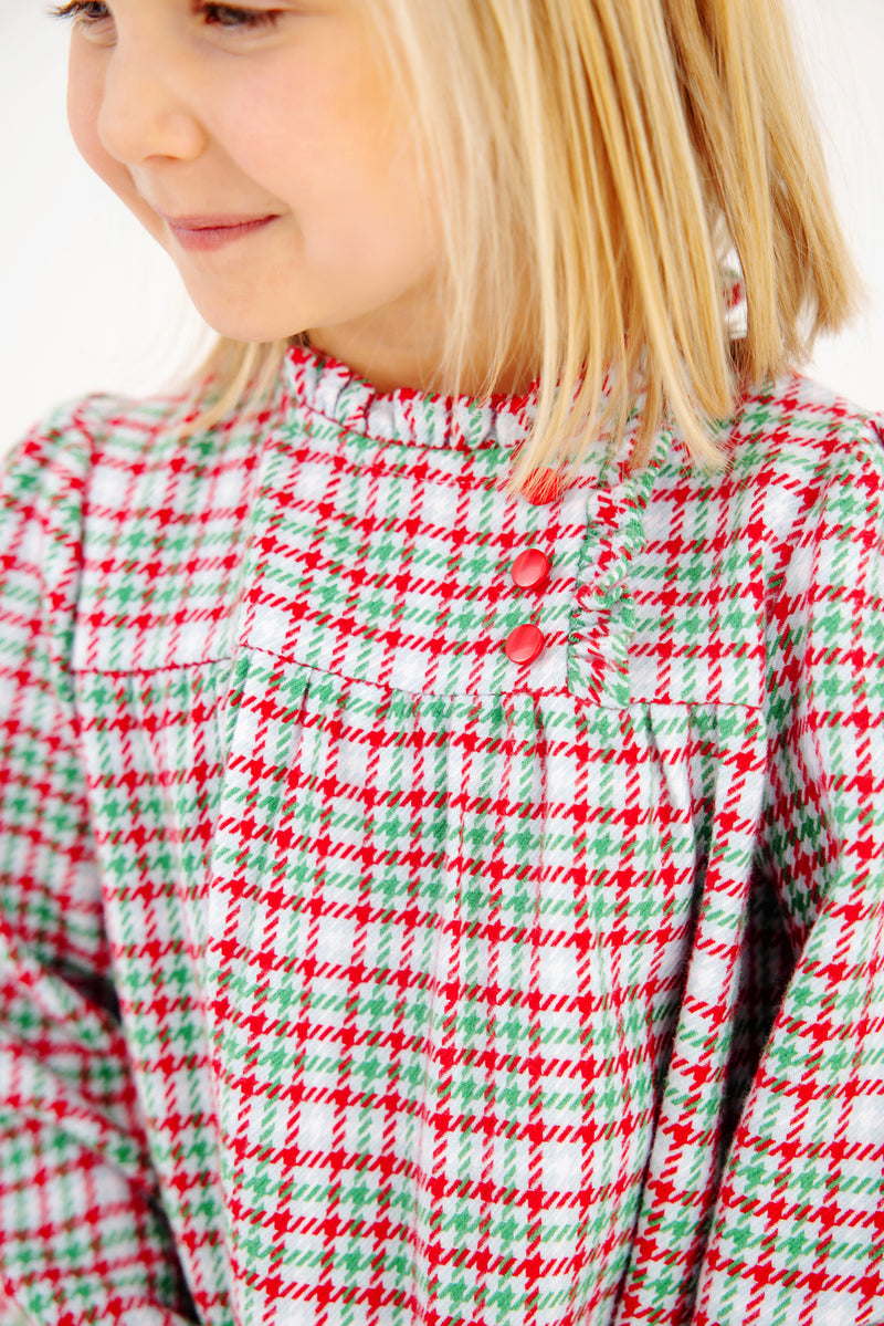 A smiling young child with blonde hair wears The Beaufort Bonnet Company’s Long Sleeve Mary Baker Day Dress in Wicker Park Plaid with Richmond Red—featuring three red buttons on the chest, it’s the perfect Christmas holiday dress.