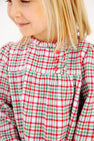 A smiling young child with blonde hair wears The Beaufort Bonnet Company’s Long Sleeve Mary Baker Day Dress in Wicker Park Plaid with Richmond Red—featuring three red buttons on the chest, it’s the perfect Christmas holiday dress.