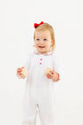A toddler wears a Long Sleeve Britt Romper - Worth Avenue White with Holly, featuring white Pima cotton, red trim, red buttons, and a small holly embroidery on the collar, while holding two wooden blocks.