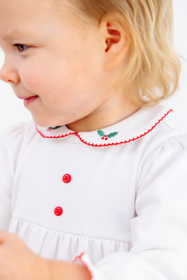 A toddler wearing a Long Sleeve Britt Romper - Worth Avenue White with Holly, a white Pima cotton romper featuring red buttons and a rounded collar embroidered with holly leaves and red berries.