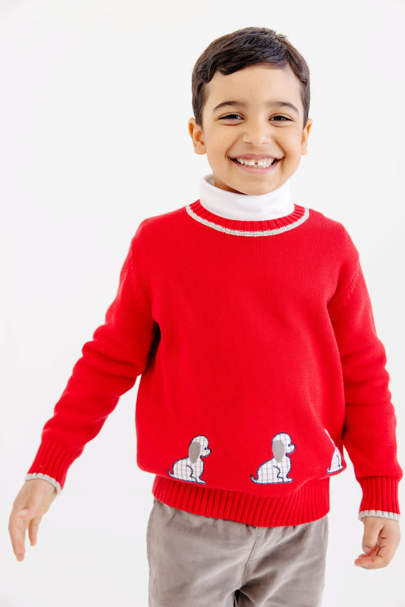A smiling boy wearing an Isaac’s Intarsia Sweater - Richmond Red with Grantley Gray and Dogs layered over a white turtleneck, featuring a red long-sleeved design with small dogs near the hem.