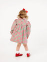 A young child with curly hair, wearing The Beaufort Bonnet Company’s Della Marie Dress in Richmond Red & Kiawah Kelly Green Gingham, white socks, red shoes, and a red bow, stands facing away from the camera against a white background.