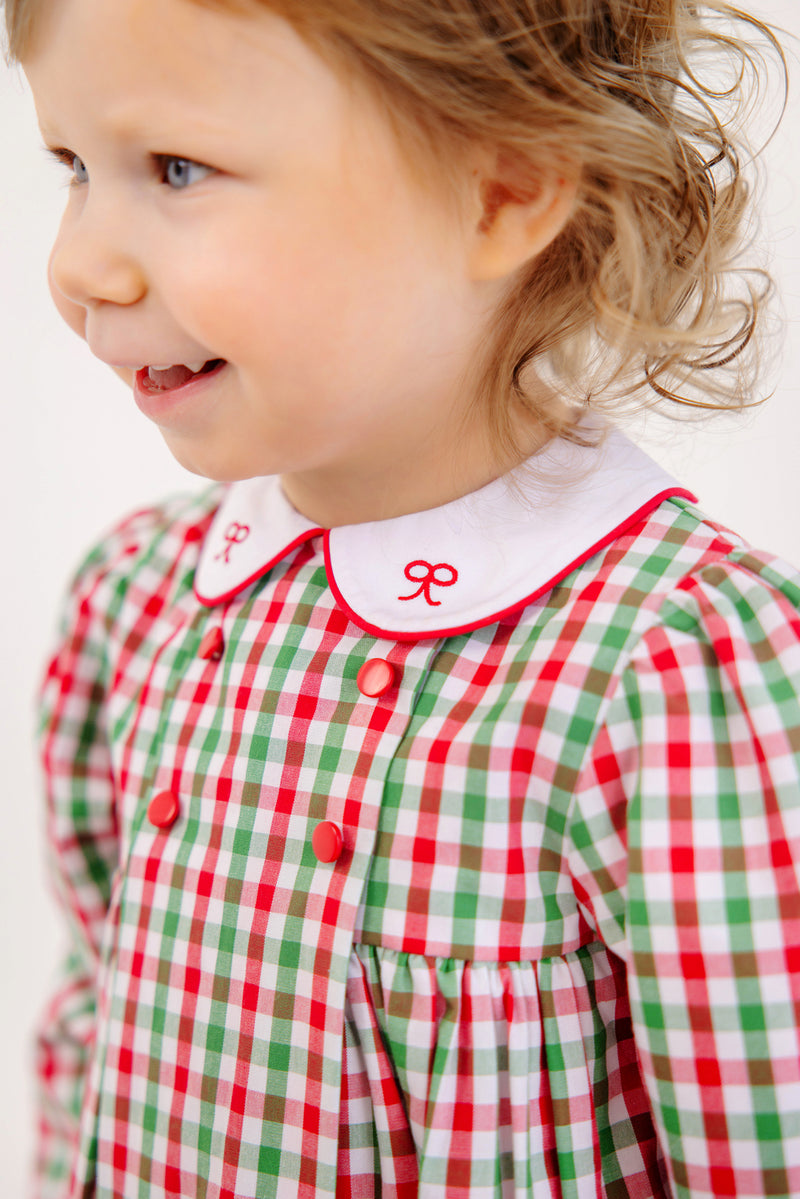 A young child smiles, wearing The Beaufort Bonnet Company’s Della Marie Dress in Richmond Red & Kiawah Kelly Green Gingham, featuring red buttons and a white collar embroidered with small red bows.