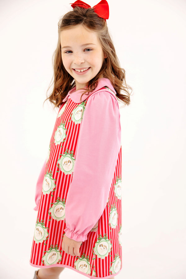 A girl wearing a Hamptons Hot Pink Annie Apron Dress - Distinguished Doggies with circular dog illustrations and a red hair bow.