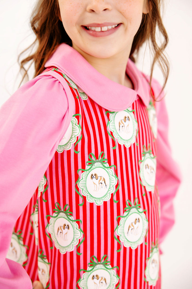A girl wearing a Maude’s Peter Pan Collar Shirt (Long Sleeve Pima) - Hamptons Hot Pink under a red and white striped jumper featuring a repeating pattern of dogs in circular frames.