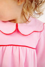 A close-up of a young child wearing The Beaufort Bonnet Company’s Long Sleeve Maude’s Day Dress in Pier Party Pink with Richmond Red, featuring a scalloped collar edged in red. Crafted from 100% Pima Cotton, the dress beautifully complements the child’s curly light brown hair as they look away from the camera.