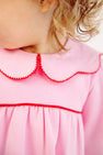 A close-up of a young child wearing The Beaufort Bonnet Company’s Long Sleeve Maude’s Day Dress in Pier Party Pink with Richmond Red, featuring a scalloped collar edged in red. Crafted from 100% Pima Cotton, the dress beautifully complements the child’s curly light brown hair as they look away from the camera.