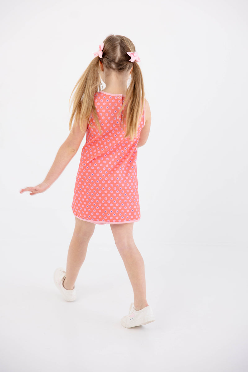 A young girl with blonde pigtails, wearing The Beaufort Bonnet Company’s Prepletic™ Taffy Tennis Dress in Mill Street Manor Mini Floral with Sandpearl Pink and white shoes, stands facing away on a white background with one arm slightly extended.