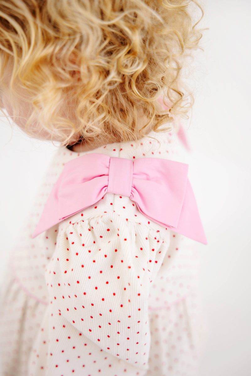 A young child with curly blonde hair wears The Beaufort Bonnet Company’s Long Sleeve Ryleigh Romper in Palmetto Pearl & Richmond Red Microdot Corduroy, styled playfully over a cozy winter outfit.