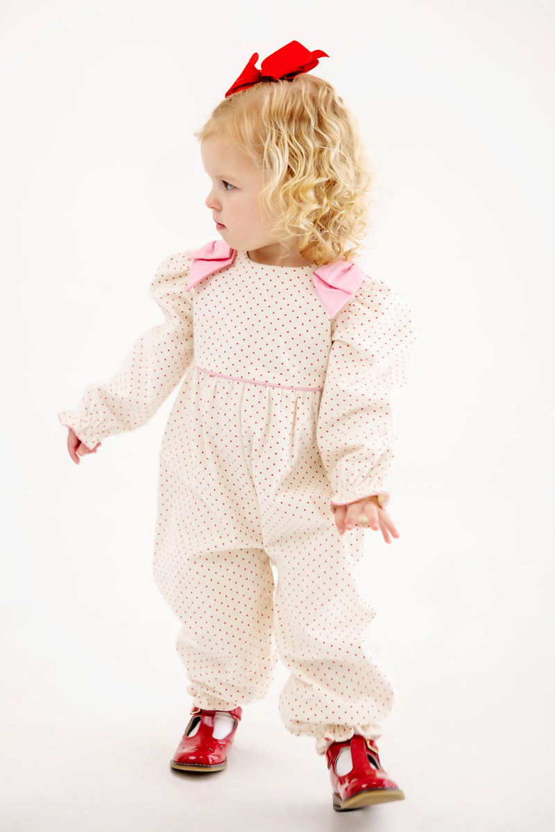 A toddler with curly blonde hair, dressed in The Beaufort Bonnet Company’s Long Sleeve Ryleigh Romper in Palmetto Pearl & Richmond Red Microdot Corduroy, featuring pink bows, red shoes, and a red bow in her hair, stands looking to the side against a plain white background.
