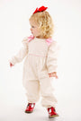 A toddler with curly blonde hair, dressed in The Beaufort Bonnet Company’s Long Sleeve Ryleigh Romper in Palmetto Pearl & Richmond Red Microdot Corduroy, featuring pink bows, red shoes, and a red bow in her hair, stands looking to the side against a plain white background.