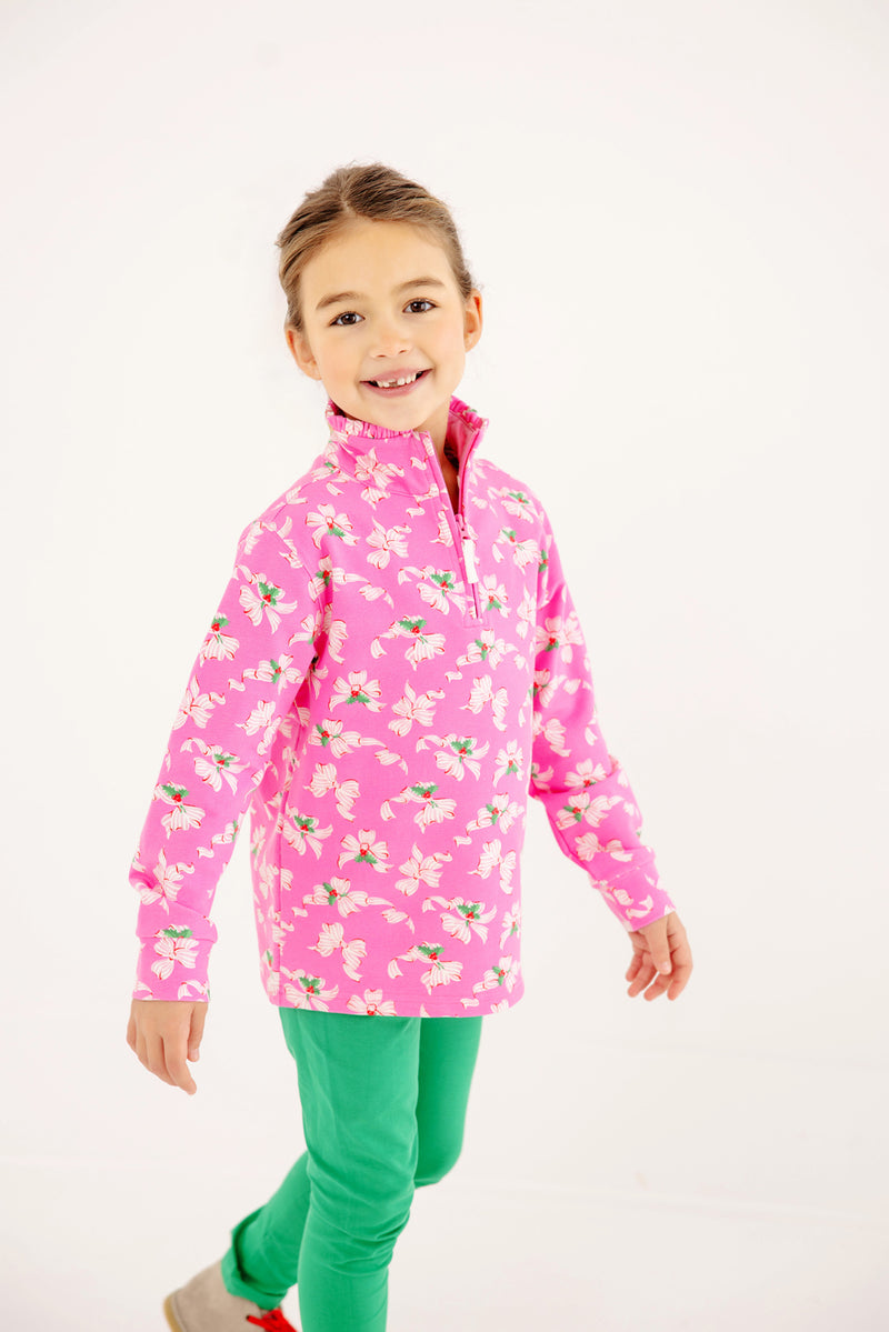 A smiling young girl with brown hair in a ponytail wears The Beaufort Bonnet Company’s Canter Collar Half Zip in the Bows of Holly print and green pants, standing against a plain white background.