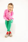 A smiling young girl with brown hair, wearing The Beaufort Bonnet Company’s Canter Collar Half Zip in the Bows of Holly print, green pants, and gray shoes with red laces, sits cross-legged on a white block against a plain white background.