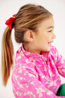 Smiling young girl with long hair in a ponytail and a red bow, wearing The Beaufort Bonnet Company’s Canter Collar Half Zip in Bows of Holly, looking to the side against a white background.