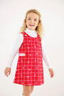 A young girl with curly blonde hair is smiling against a plain, light background, wearing The Beaufort Bonnet Company’s Janie Jumper in Torrington Tartan with Pier Party Pink over a white turtleneck.