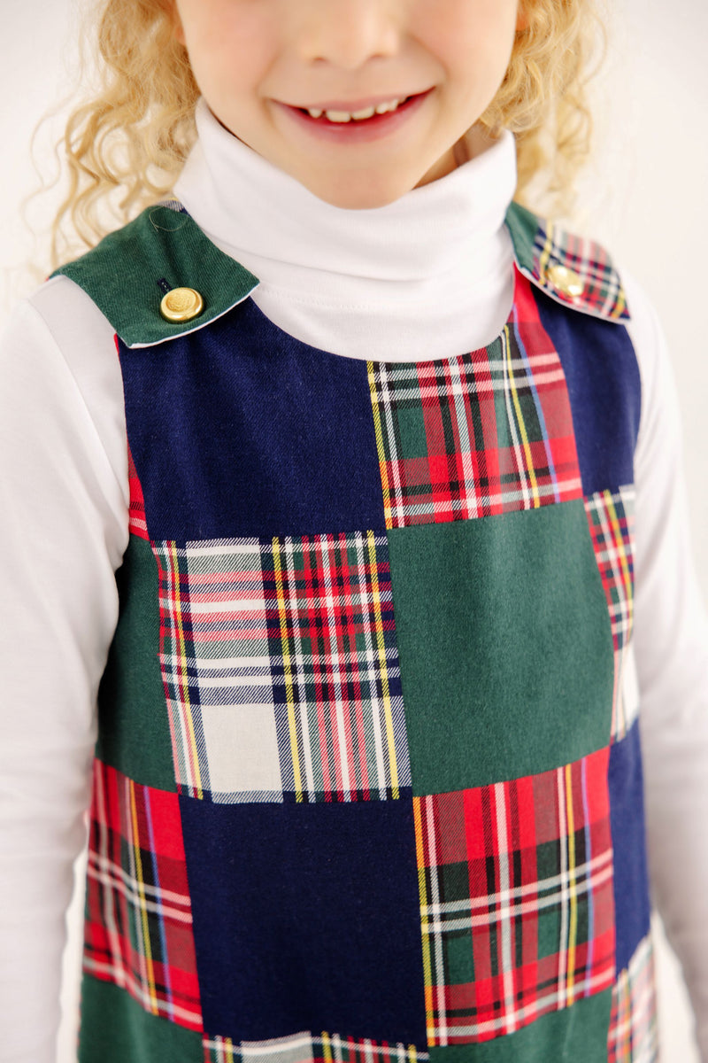 A child wears a Mercer Island Madras Juliet Jumper in a patchwork of solid green, navy blue, and colorful plaid fabric, styled with gold buttons at the shoulders over a white turtleneck.