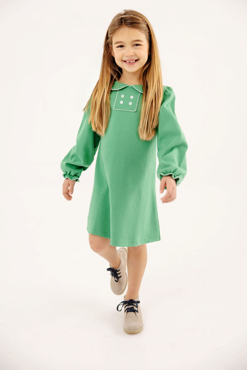 A young girl wearing a The Beaufort Bonnet Company Long Sleeve Banks Button Dress - Kiawah Kelly Green with Worth Avenue White, featuring white button details, a white-trimmed collar, and beige shoes.
