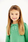 A young girl smiles wearing a The Beaufort Bonnet Company Long Sleeve Banks Button Dress - Kiawah Kelly Green with Worth Avenue White, featuring a white-trimmed collar and a decorative square panel with four white buttons.