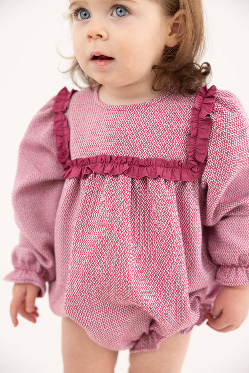 A toddler with light brown hair and blue eyes wears The Beaufort Bonnet Company’s Debbie Day Bubble in Begley Berry Tweed, featuring textured pink fabric with darker pink ruffles on the chest and shoulders, standing against a plain white background.