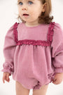 A toddler with light brown hair and blue eyes wears The Beaufort Bonnet Company’s Debbie Day Bubble in Begley Berry Tweed, featuring textured pink fabric with darker pink ruffles on the chest and shoulders, standing against a plain white background.