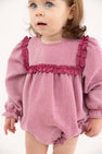 A toddler with light brown hair and blue eyes wears The Beaufort Bonnet Company’s Debbie Day Bubble in Begley Berry Tweed, featuring textured pink fabric with darker pink ruffles on the chest and shoulders, standing against a plain white background.