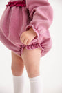 Dressed in The Beaufort Bonnet Company’s Debbie Day Bubble in Begley Berry Tweed, a toddler stands against a plain white background—this textured romper with ruffled edges, paired with white knee-high socks, makes for a charming baby girl outfit perfect for autumn.