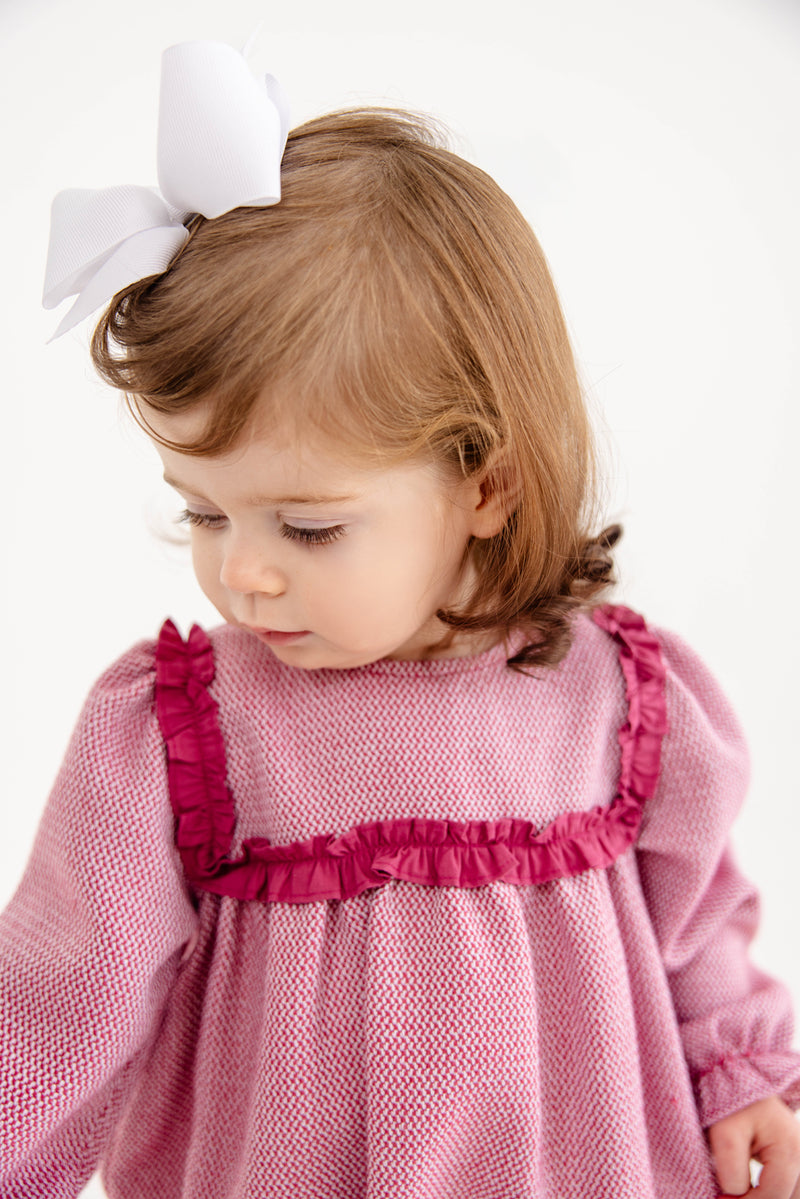 A young child with light brown hair and a white bow looks down, wearing The Beaufort Bonnet Company’s Debbie Day Bubble in Begley Berry Tweed—a charming pink outfit with darker pink ruffle trim—styled as an adorable baby girl ensemble against a plain white background.