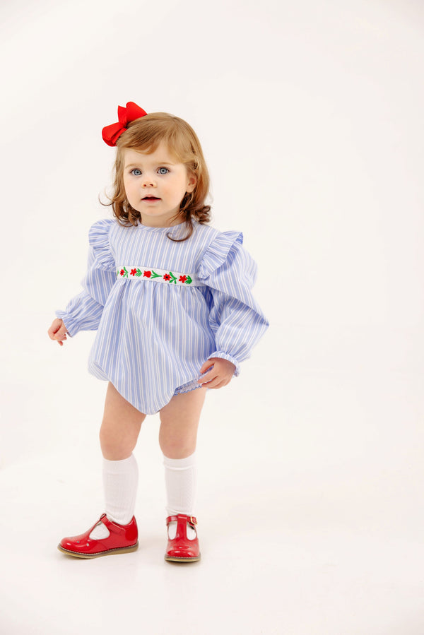 A toddler wears The Beaufort Bonnet Company Long Sleeve Ruehling Ruffle Bubble - Park City Periwinkle Ticking Stripe & Worth Avenue White, a blue and white striped long sleeve romper with floral embroidery, white socks, and red shoes.