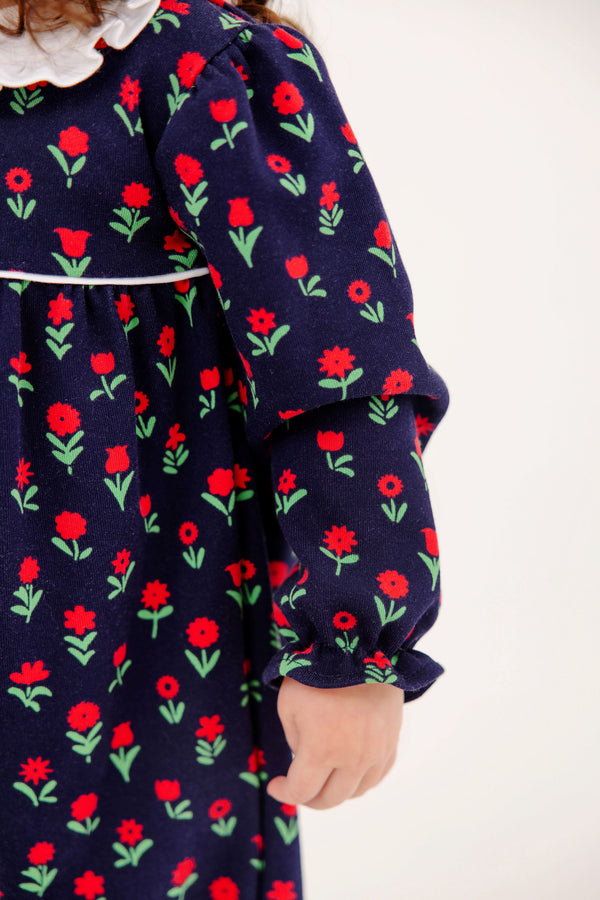 Maude’s Ruffle Romper - Prescott Farms Flowers with Worth Avenue White is a long-sleeved navy blue baby girl ruffle romper featuring a repeating pattern of red flowers with green stems and a white ruffled collar.