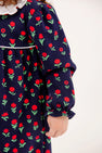 Maude’s Ruffle Romper - Prescott Farms Flowers with Worth Avenue White is a long-sleeved navy blue baby girl ruffle romper featuring a repeating pattern of red flowers with green stems and a white ruffled collar.