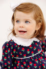 A toddler wearing Maude’s Ruffle Romper - Prescott Farms Flowers with Worth Avenue White, featuring a navy blue floral print with red tulips, a white ruffled collar, and a white bow in her hair.